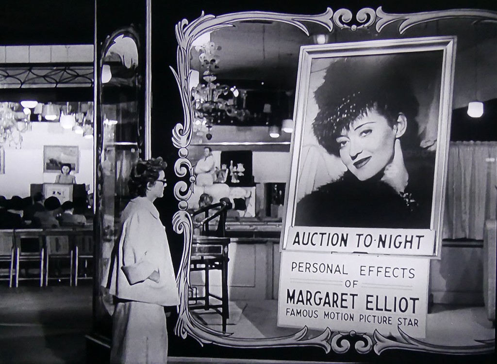 First scene of black and white movie, "The Star", 1952. Night. Bette Davis, in sunglasses, walking on sidewalk, stops in front of store window. Right hand in pocket of cloth jacket. While auction can be heard through open door to her left, looks at at large photo of earlier self above sign: "Auction To-Night. Personal Effects of Margaret Elliot, Famous Motion Picture Star". Photo is of the real Bette Davis, as Judith Traherne, in "Dark Victory", 1939. Nominated for Best Actress Oscar.